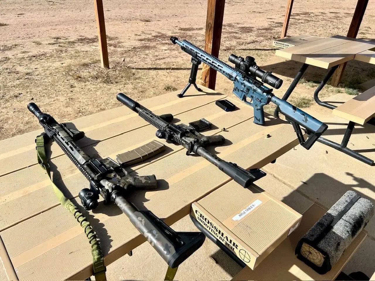Three rifles with scopes and accessories on a wooden table at an outdoor shooting range.