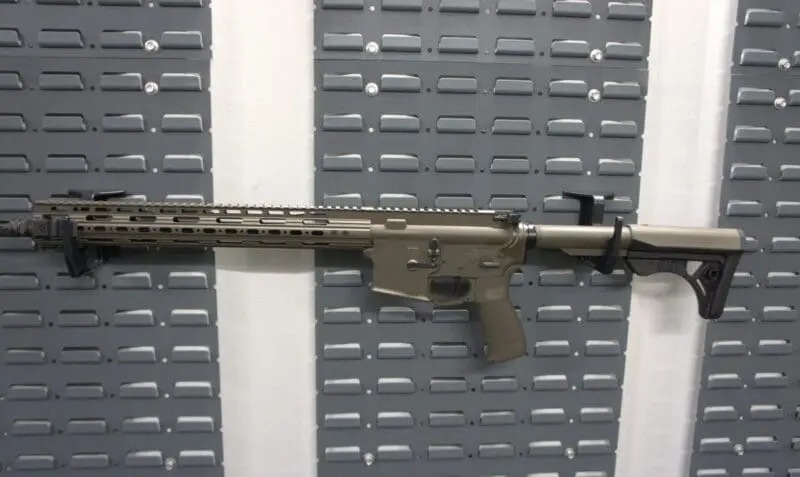 A modern tactical rifle with a tan finish against a gray wall.